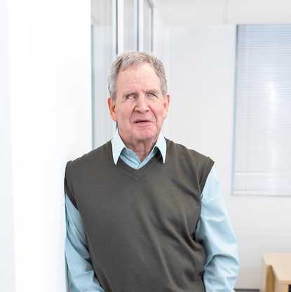 Mike Brace, Chair of BWBF, photographed in 2024. He is pictured leaning against a wall in an office and holding the lead for his guide dog.