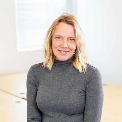 Jenny Cameron, Head of Communications, Marketing and Income for BWBF, sits on a table smiling to camera. She has blonde, wavy, shoulder-length hair and is wearing a grey top.