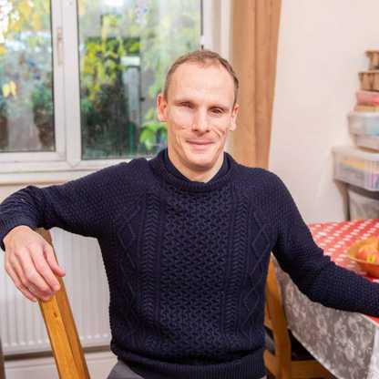 Jason, a user of BWBF's apps, sits at a table with a red checked table cloth. He is smiling and wearing a dark jumper.
